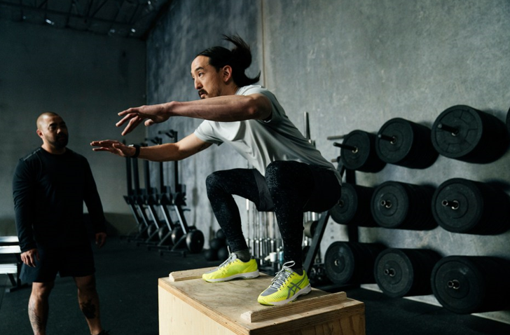 A personal trainer watching Steve Aoki perform high intensity interval training (HIIT) on top of a wooden box.  