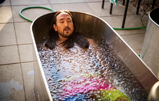 Steve Aoki submerged in an ice bath from the neck down, wearing rainbow-colored shorts. 